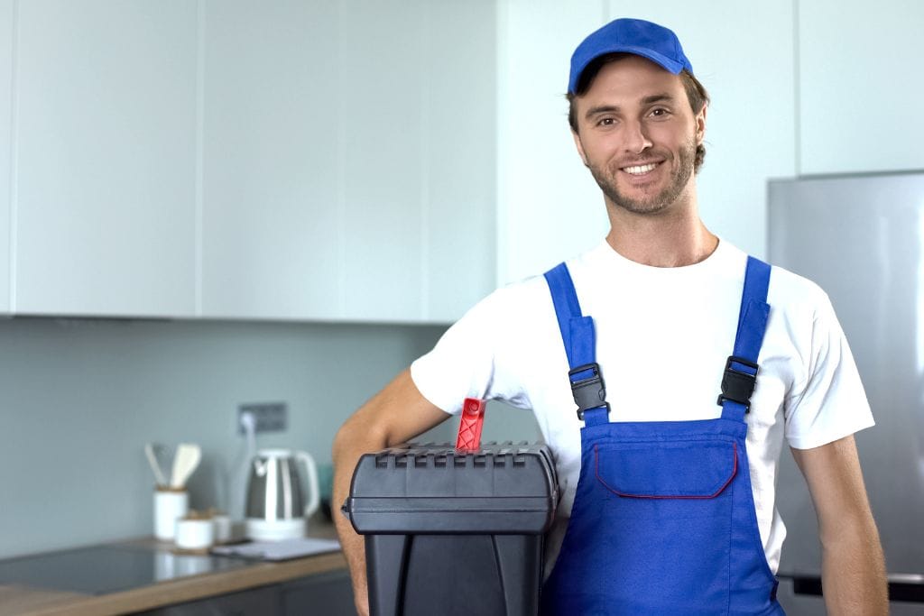 A man in overalls holding a trash can in front of a kitchen.